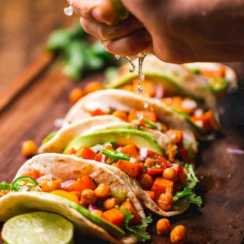 A person fills tacos with chopped vegetables and beans, squeezing ingredients as sauce drips onto the taco shells.