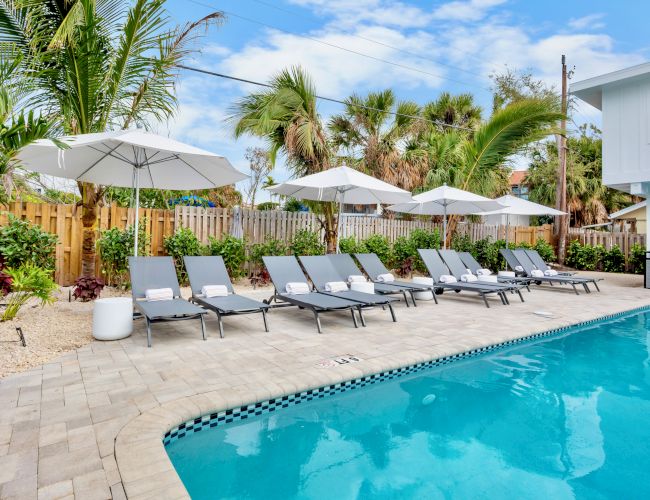 A sunny poolside scene with a row of lounge chairs under white umbrellas, tropical plants, and a bright blue pool next to a white building.