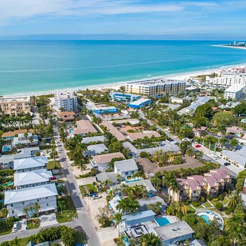 Aerial view of a coastal town with palm trees, row houses, and low-rise buildings along a curved white-sand beach and turquoise sea, sunny and inviting.