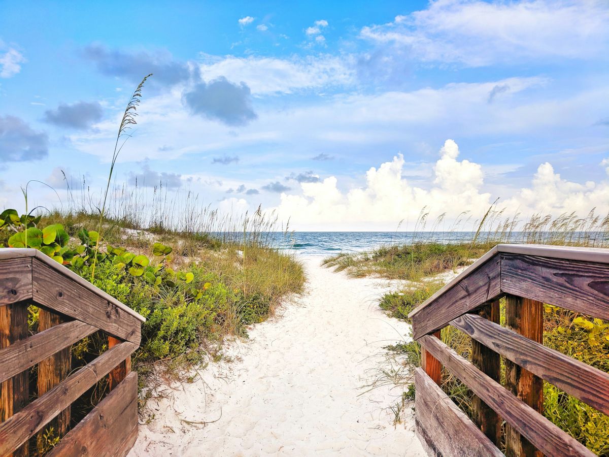 A sandy path between wooden railings leads to a bright beach with blue skies, fluffy clouds, and ocean waves in the distance.