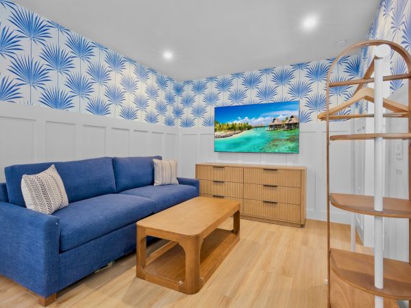 Cozy living room with blue sofa, wood coffee table, dresser, wall-mounted TV, bright palm-pattern wallpaper, and a curved wooden shelving unit.