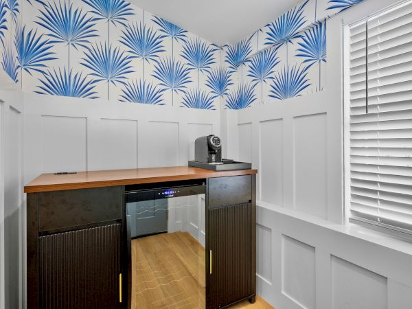 A small home bar area with dark wood cabinets, a compact fridge, and a wooden countertop, set against white paneled wainscoting and blue palm-pattern wallpaper. End.