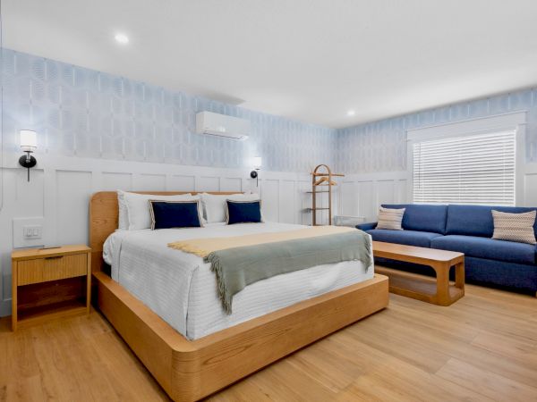 A modern hotel room with a wooden low-profile bed, light bedding, two nightstands, a blue sofa, a wooden coffee table, and soft lighting.