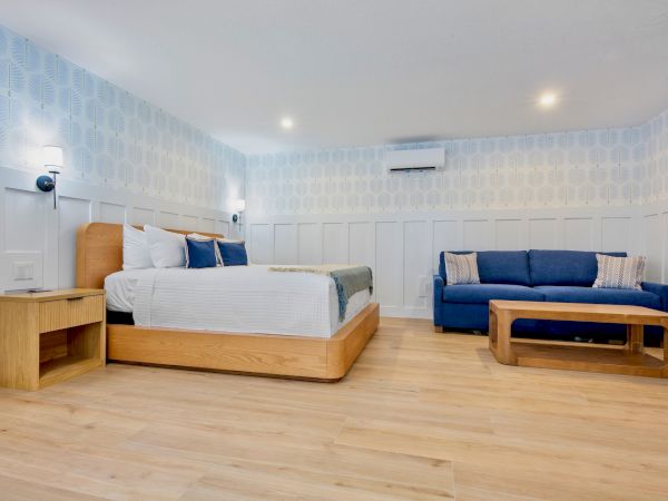 A bright, modern hotel room with a wooden bed and side table, a blue sofa, wooden coffee table, light walls, and warm hardwood floors.