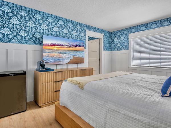 A bright hotel-style bedroom with blue damask wallpaper, white wainscoting, a wooden dresser, a TV on top, a small fridge, and a bed with white linens and blue accents, under a window.
