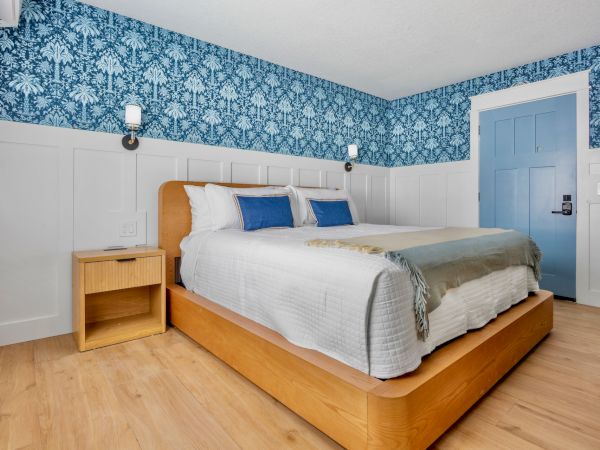 A wooden platform bed with white bedding, blue throw pillows, two small nightstands, blue floral wallpaper, and a light wood floor.