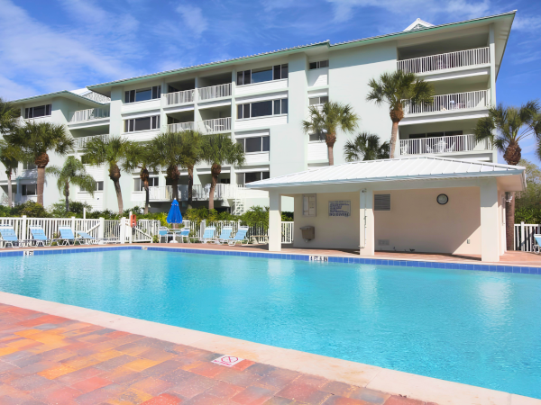 A sunny resort with a large swimming pool, palm trees, and a white multi-story building in the background, blue skies overhead, lounge chairs along the pool.